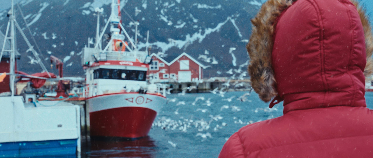 A figure in a red coat faces fishing boats in a winter harbour